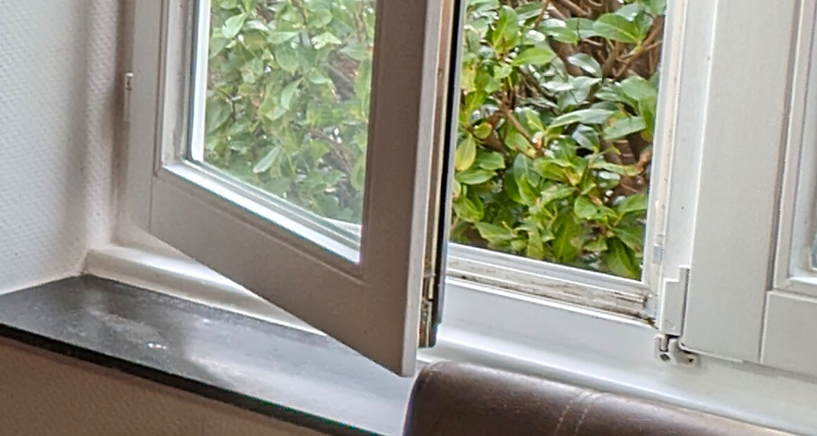 Offenes Fenster von innen mit Blick auf grüne Blätter außerhalb; Fensterbank sichtbar.