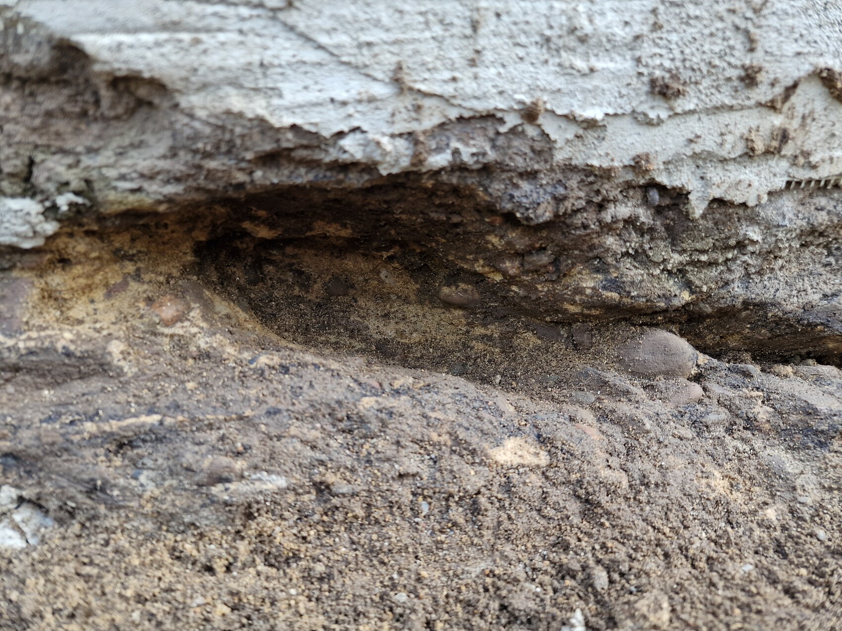 Nahaufnahme eines dunklen Erdlochs unter einer bröckelnden Wand mit Sand und Kies.