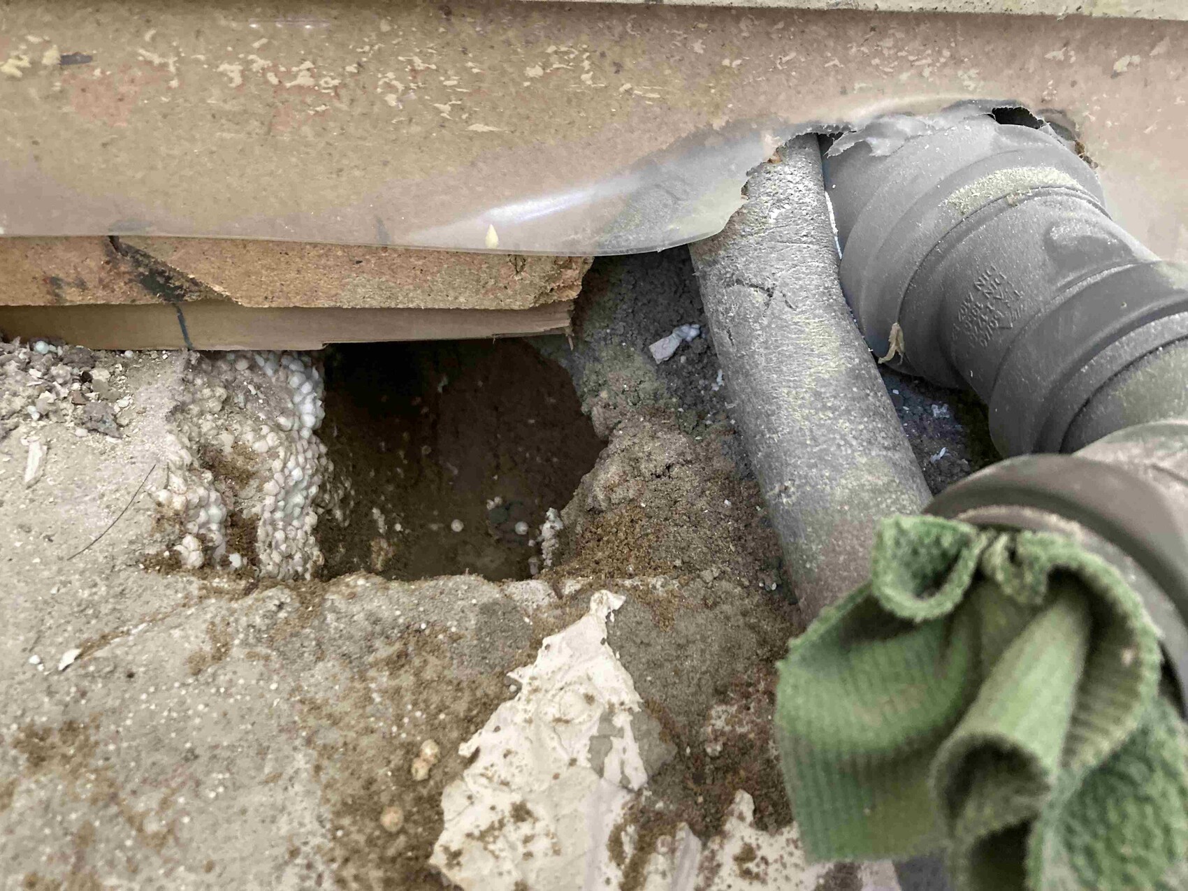 Offenes Loch im Boden mit grauem flexiblen Schlauch, Dreck, Schaum und grünem Tuch.