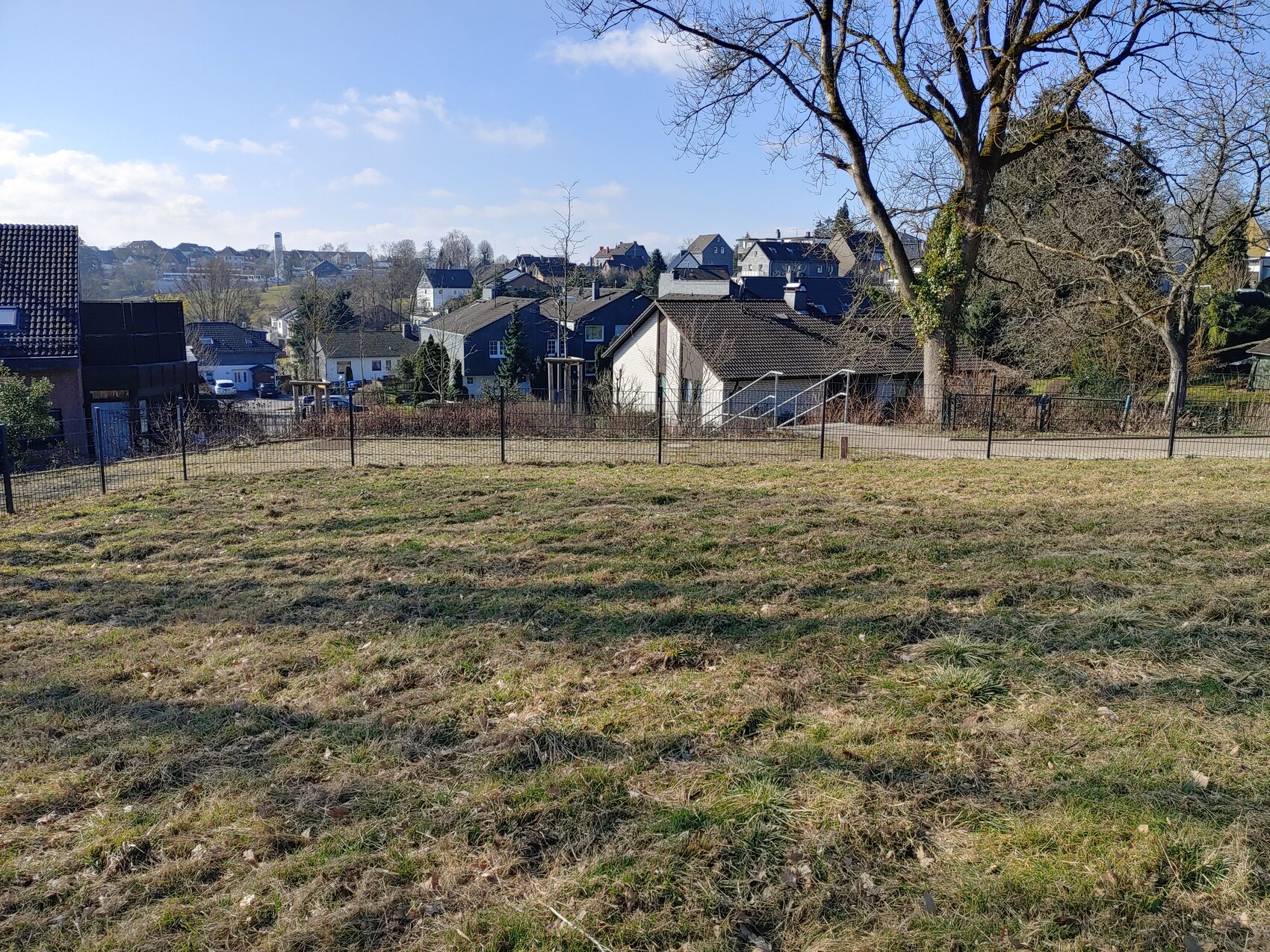 Offenes Grasfeld mit Zaun, dahinter Vorstadthäuser, kahle Bäume und blauer Himmel