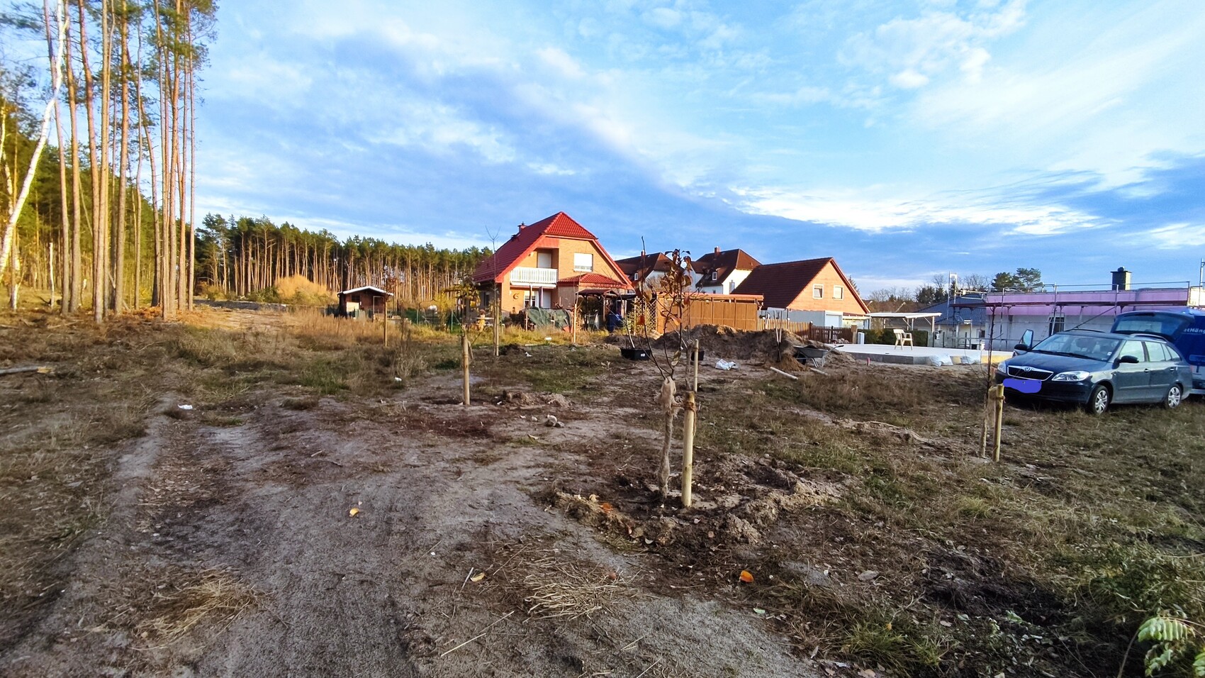 Baugebiet mit roten Ziegeldächern, Bauarbeiten, linke Baumreihe, rechts geparkte Autos.