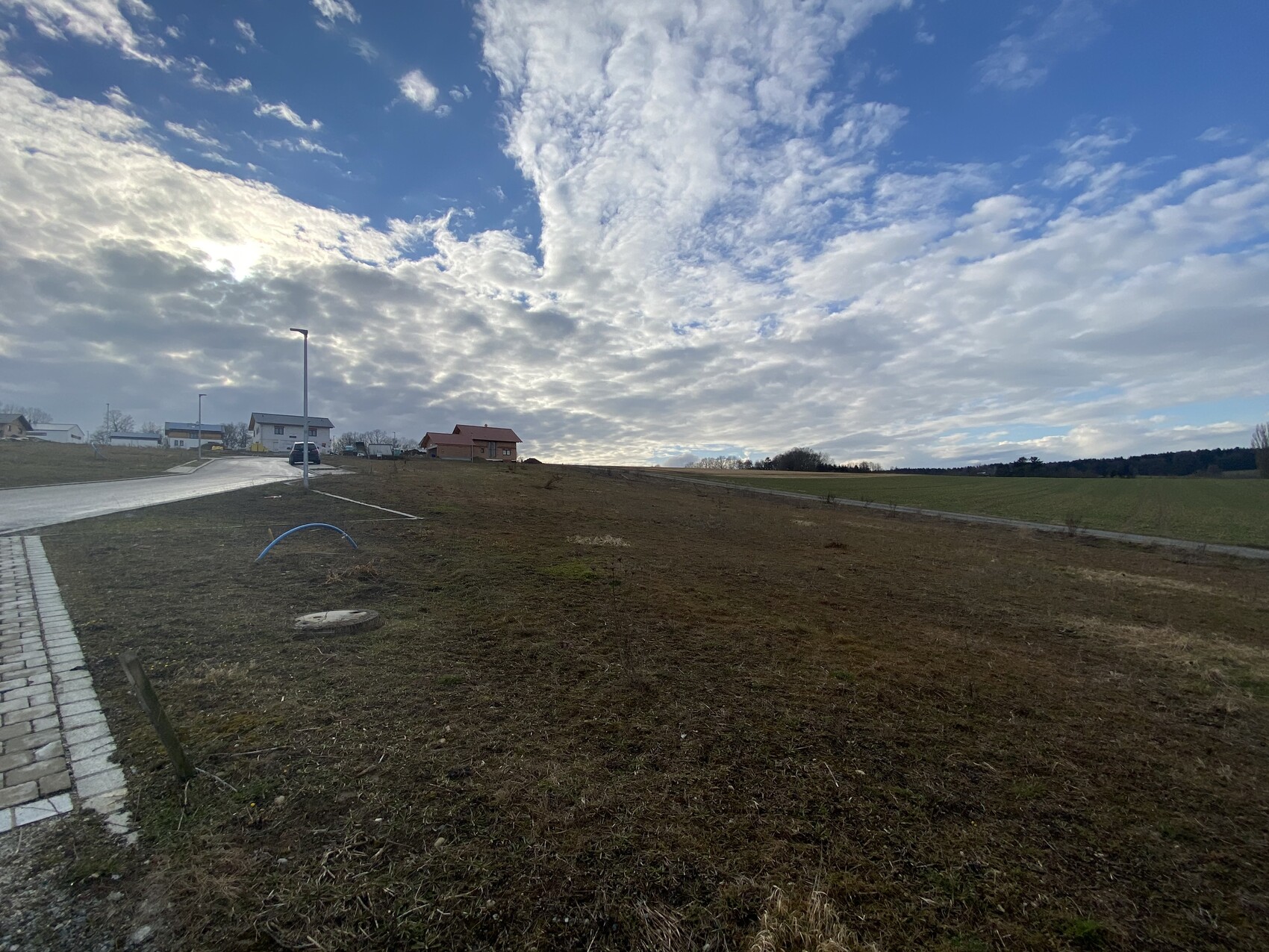 Offenes Feld mit Gehsteig links, blauer Rohrbogen, Häuser in der Ferne unter wolkigem Himmel.