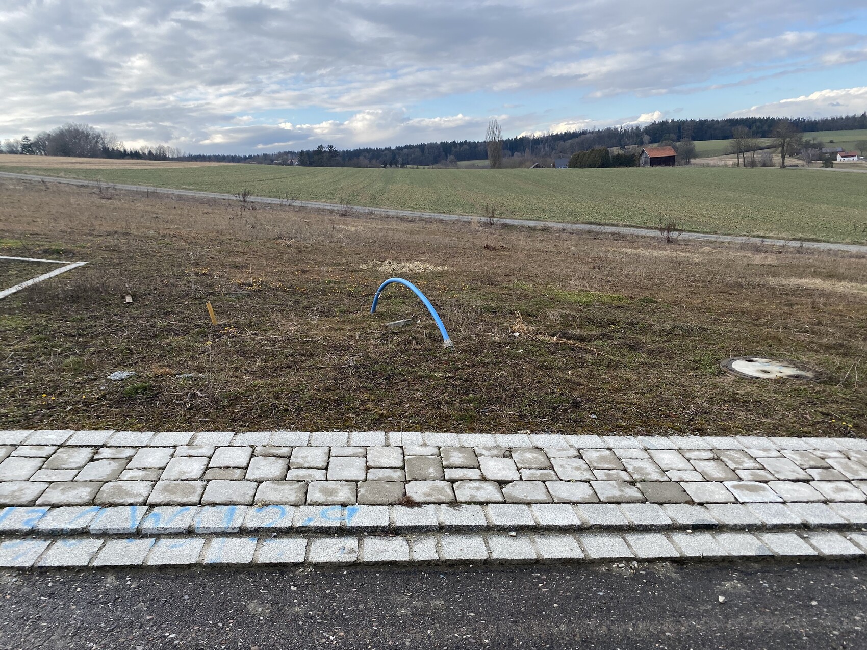 Brachland mit blauem Rohrbogen, runder Deckel rechts, gepflasterter Bordstein vorn, Felder dahinter.