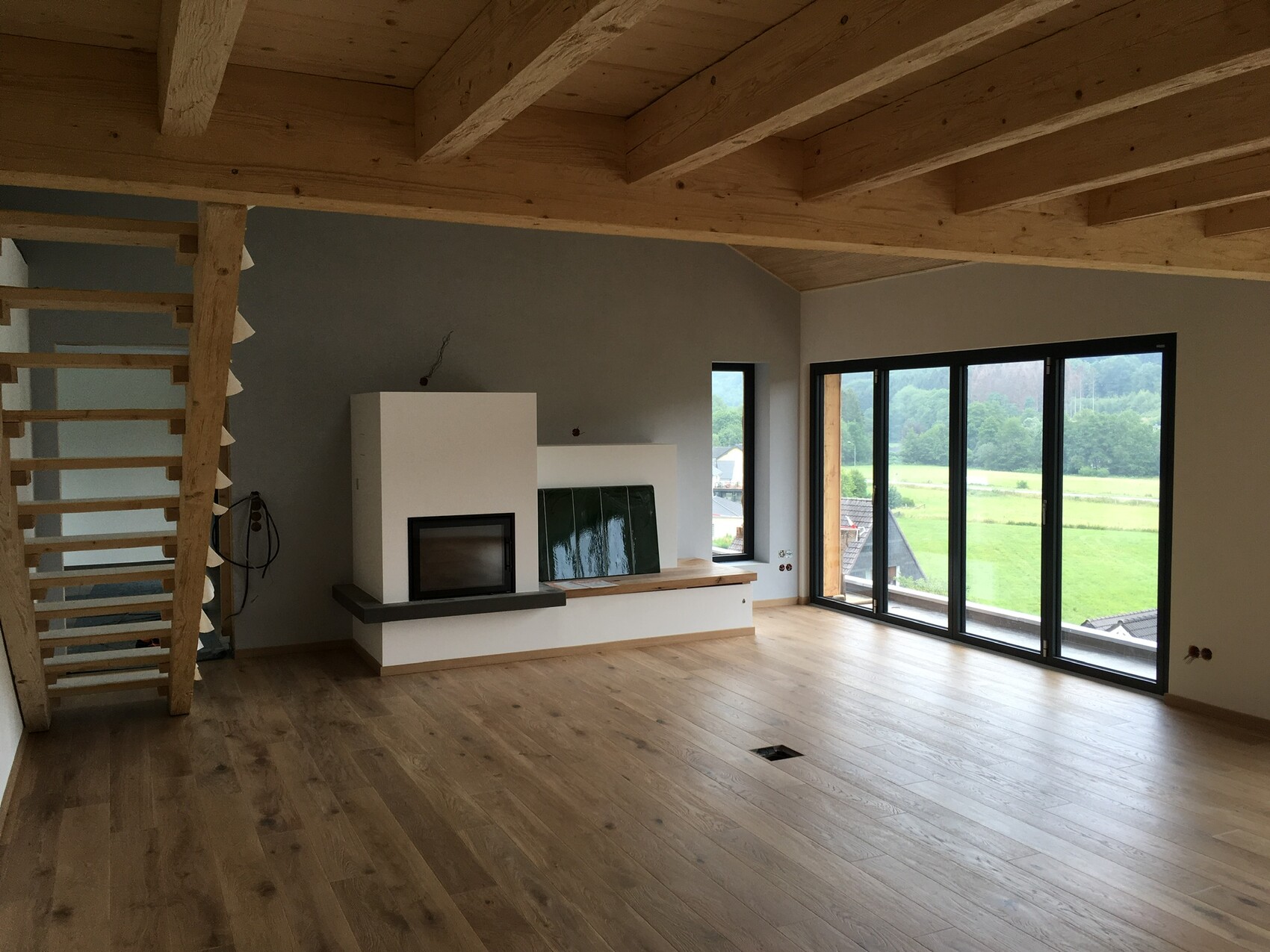 Helles Wohnzimmer mit Holzboden, sichtbaren Balken, Kamin in Weiß, Glasfront mit Blick ins Grüne.