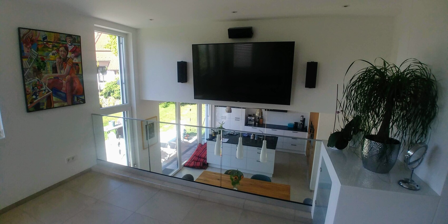 Loft-Wohnzimmer: TV-Wand, Glasgeländer, offene Küche unten, Pflanze und farbiges Gemälde.