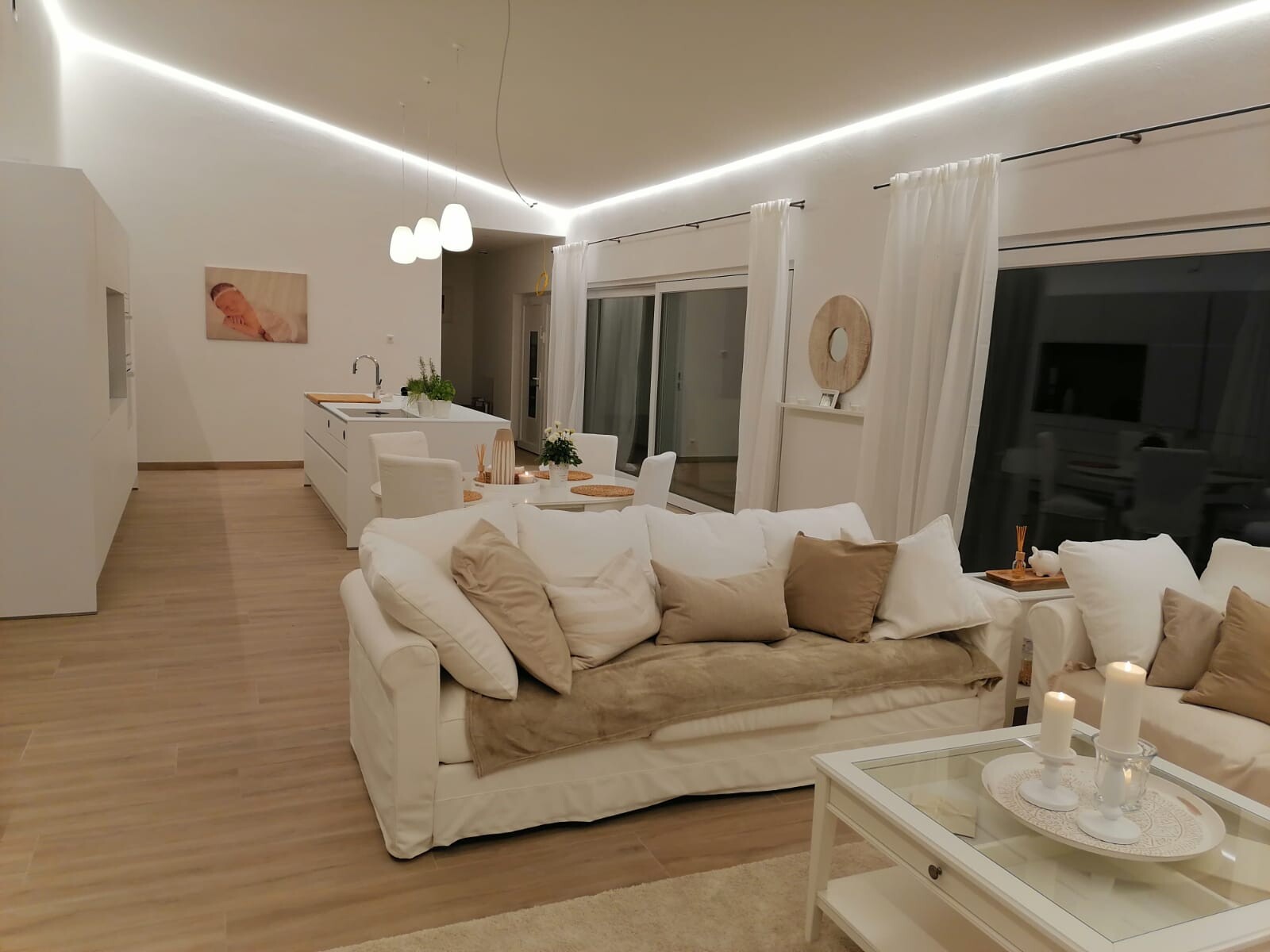 Offener Wohnbereich mit weißem Sofa, beige Kissen, Glas-Couchtisch und Küche im Hintergrund.