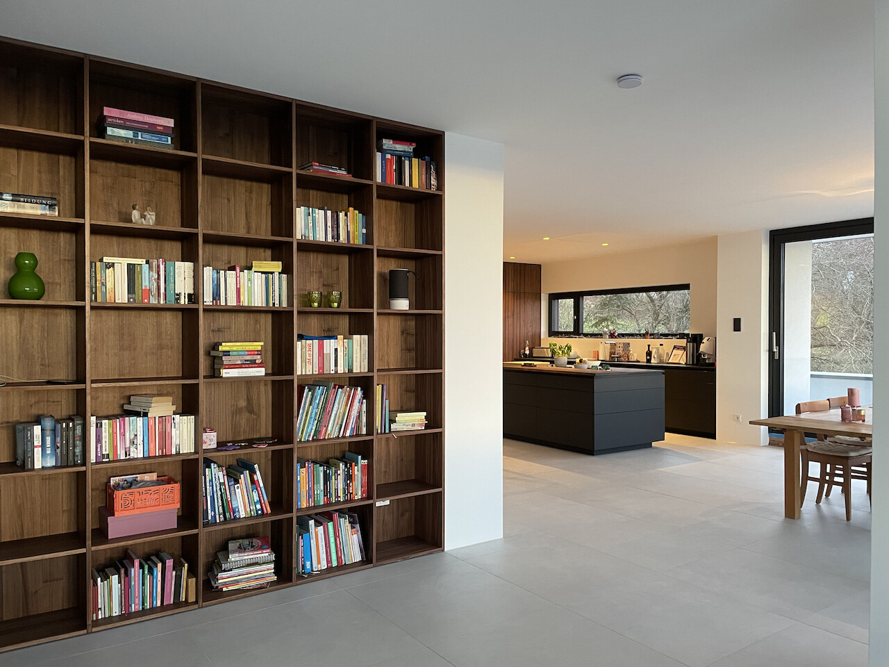 Große Holzregalwand mit vielen Büchern links; offene Küche mit Insel und Esstisch rechts.