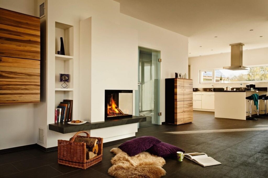 Heller offener Wohnraum mit Kamin, Kücheninsel und Sitzkissen auf dunklem Boden.
