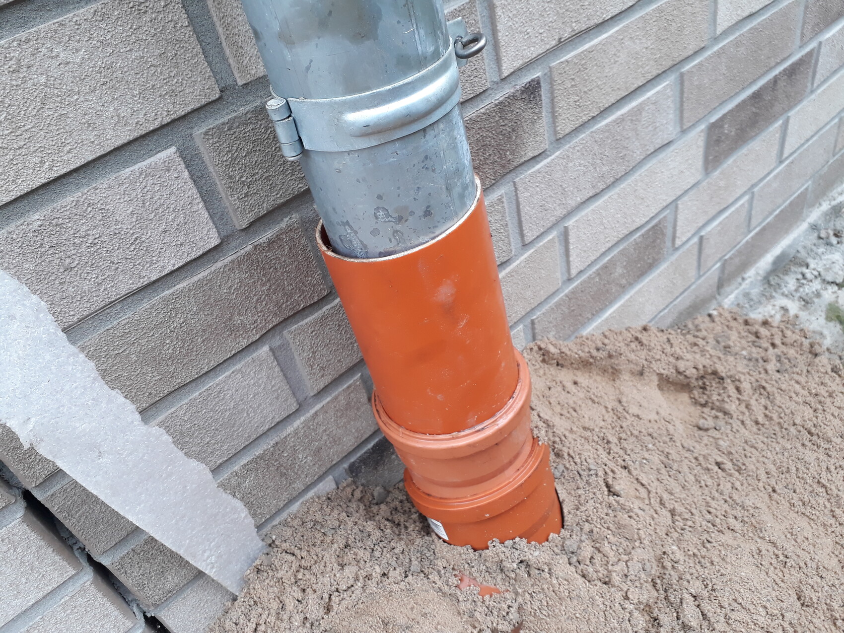Graues Metall-Abflussrohr neben einer orangefarbenen PVC-Schutzhülse am Baugrund einer Backsteinwand.