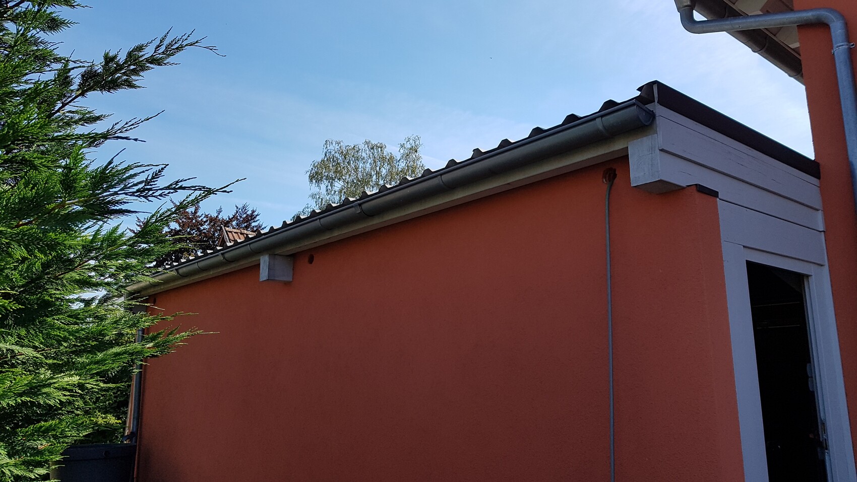 Orangefarbene Gebäudewand mit weißer Holzverkleidung, offene Tür, blauer Himmel, Bäume links.
