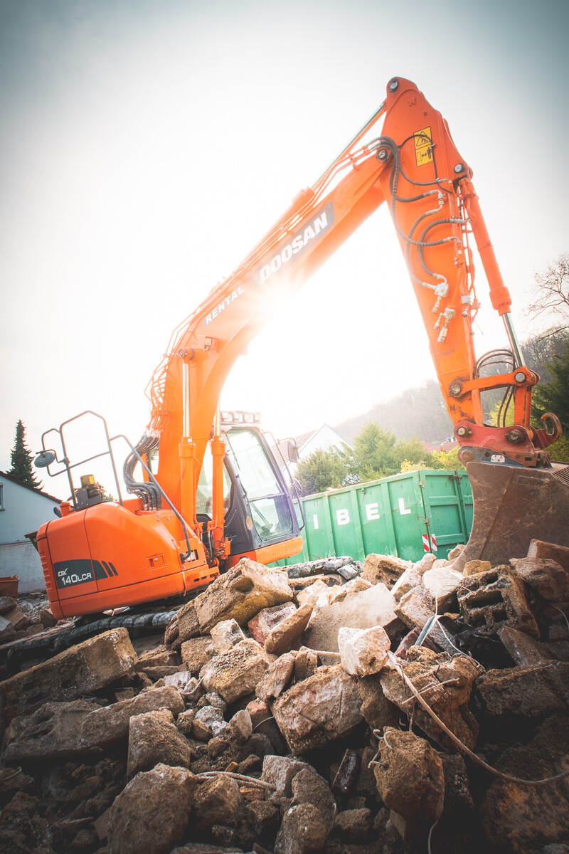 Oranger Doosan-Bagger beim Abriss auf einem Haufen Steine, grüne Container im Hintergrund