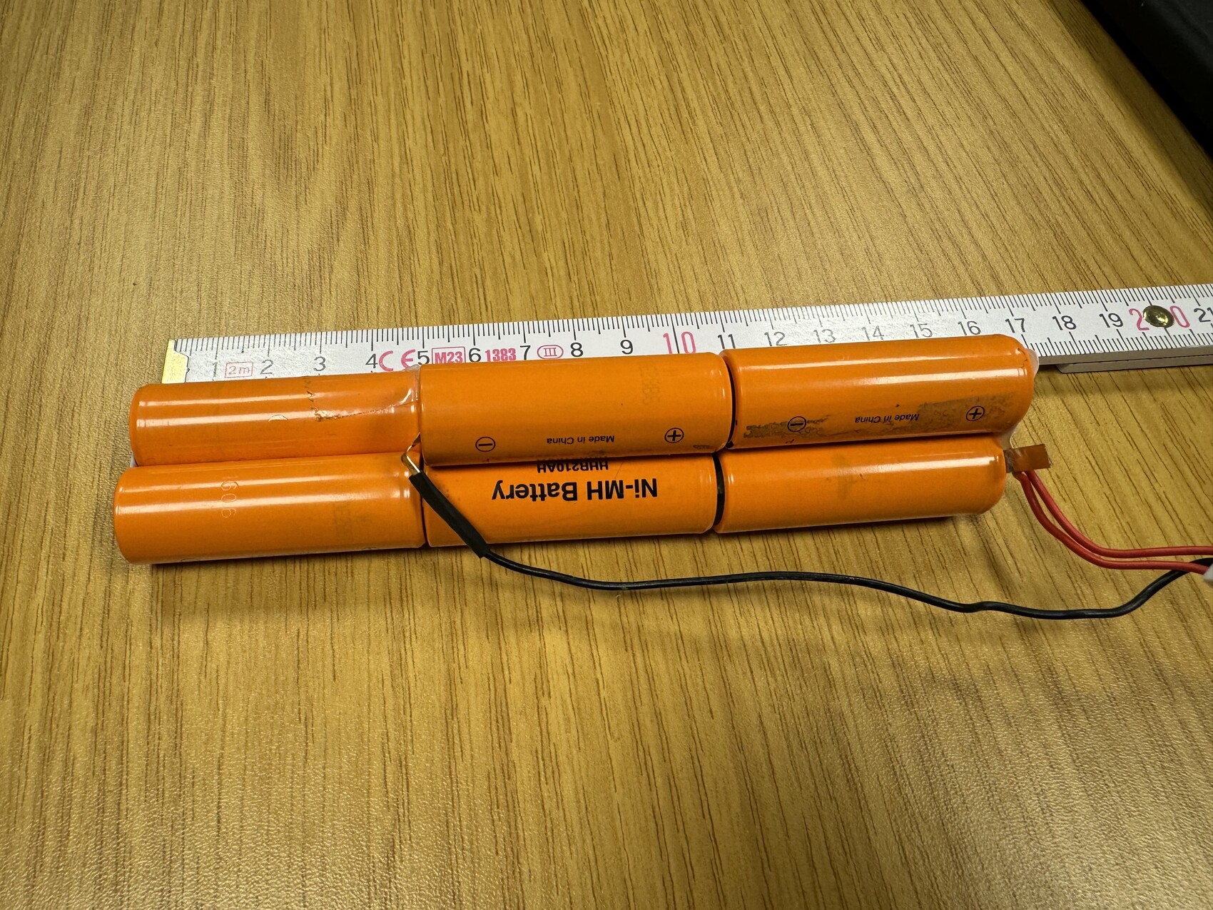 Mehrfachbatterie aus orange NiMH-Zellen mit Kabeln, neben Maßband auf Holztisch.