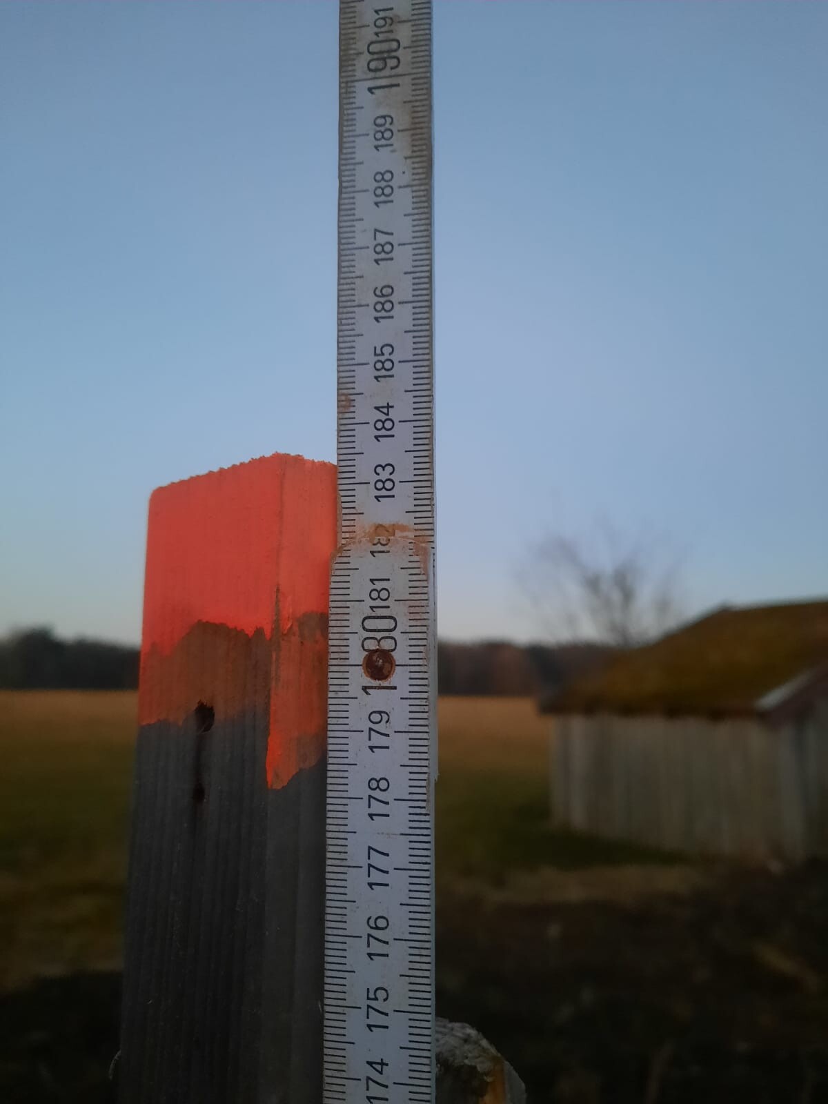Holzpfosten mit roter Spitze, daneben ein senkrecht angebrachtes Messband; ländliche Szene.