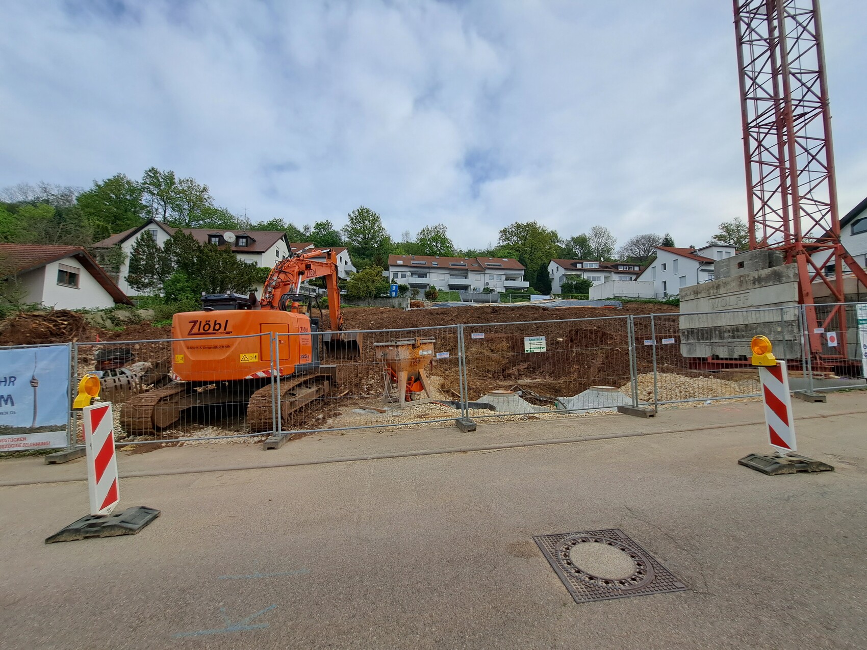 Baustelle mit orangeem Kettenbagger hinter Bauzaun, Wohnhäuser im Hintergrund.