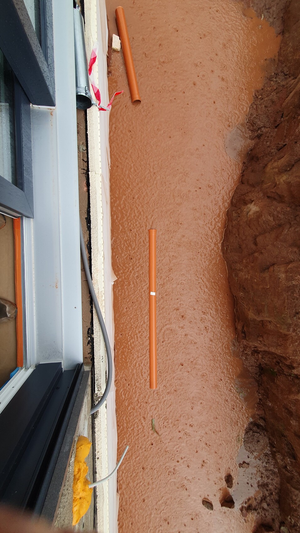 Orange PVC-Rohre liegen in einer Schlammgrube neben einem Fensterausschnitt bei Bauarbeiten.