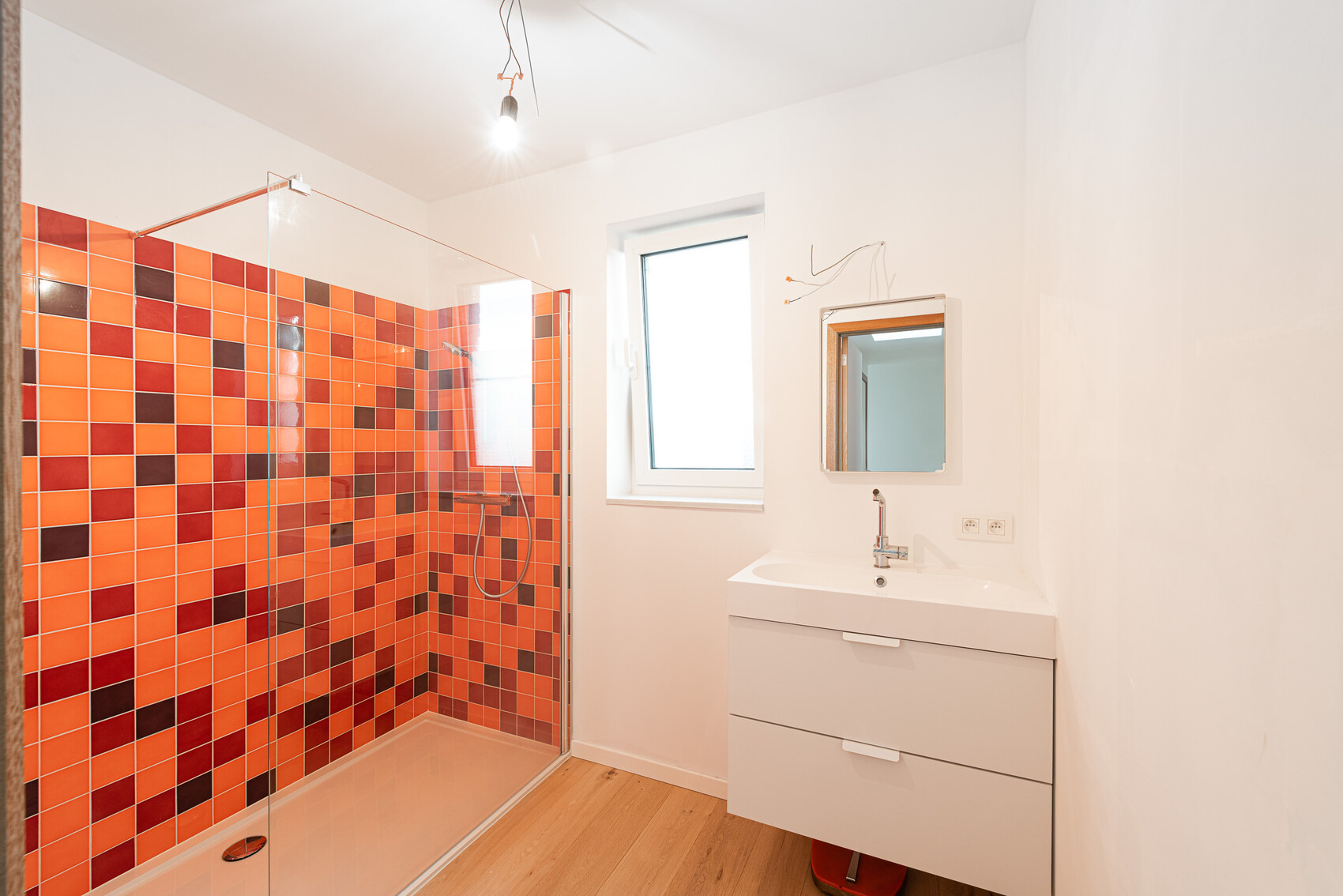 Badezimmer mit Glasdusche, orange-rote Fliesen, weißem Waschtisch, Fenster und Spiegel.