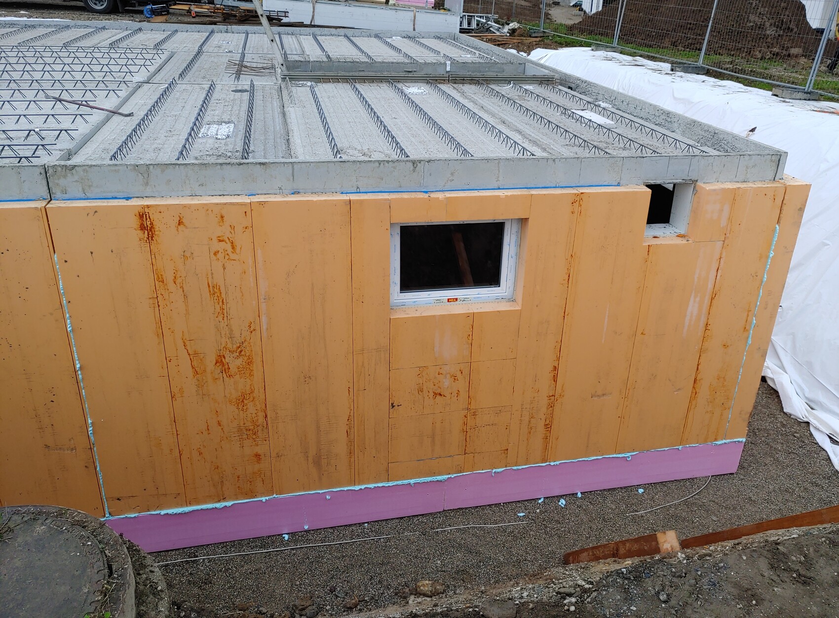 Baustelle: orangeverkleidete Außenwand mit kleinem Fenster, Bewehrung im Betonboden, pinke Dämmung unten