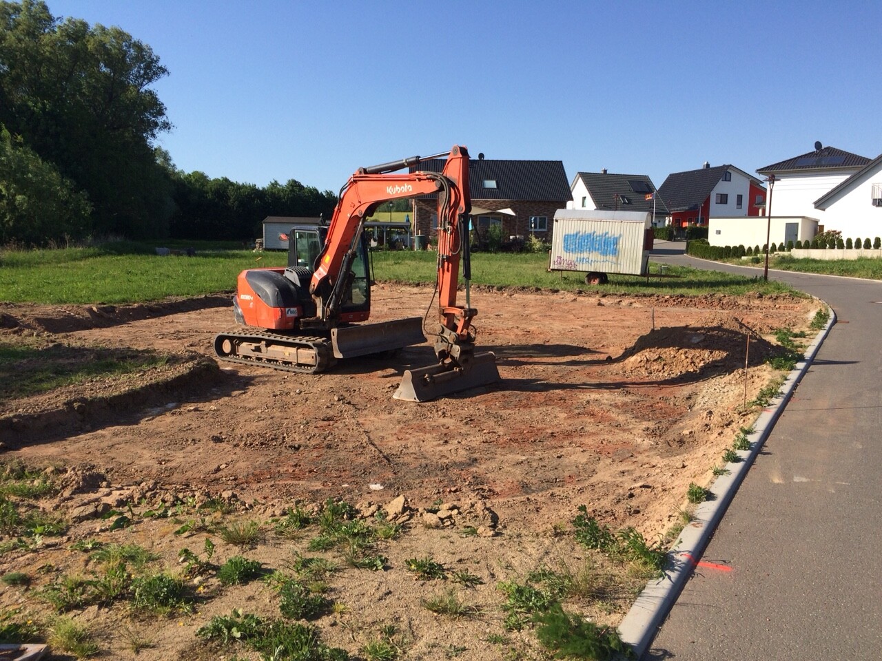 Kleine Baugrube mit orangefarbigem Bagger auf Baustelle, Wohnhäuser im Hintergrund.