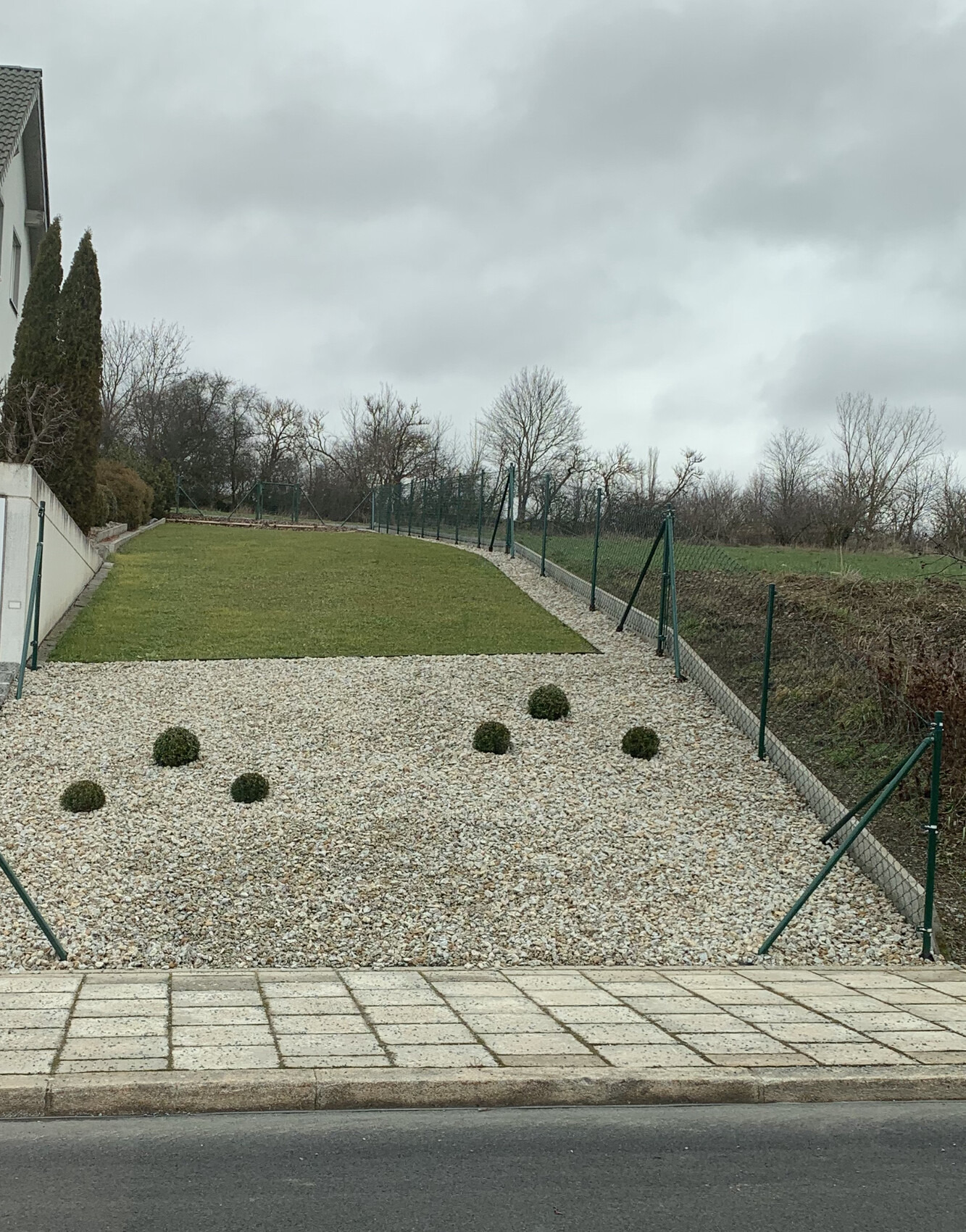 Garten mit Kiesfläche, kleinen runden Sträuchern, Grasfläche und Zaun am Hang.