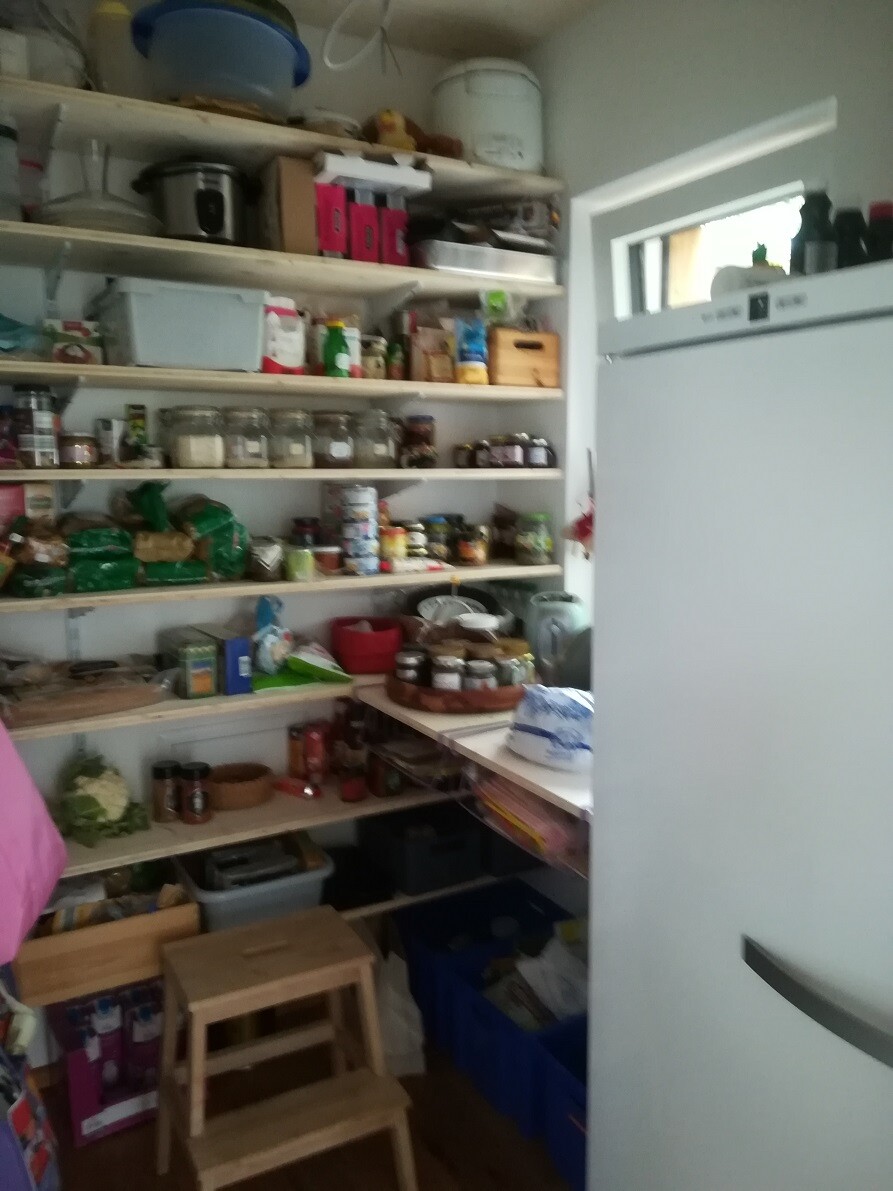 Vorratskammer mit offenen Regalen voller Gläser, Dosen und Vorratsbehälter; Kühlschrank rechts.