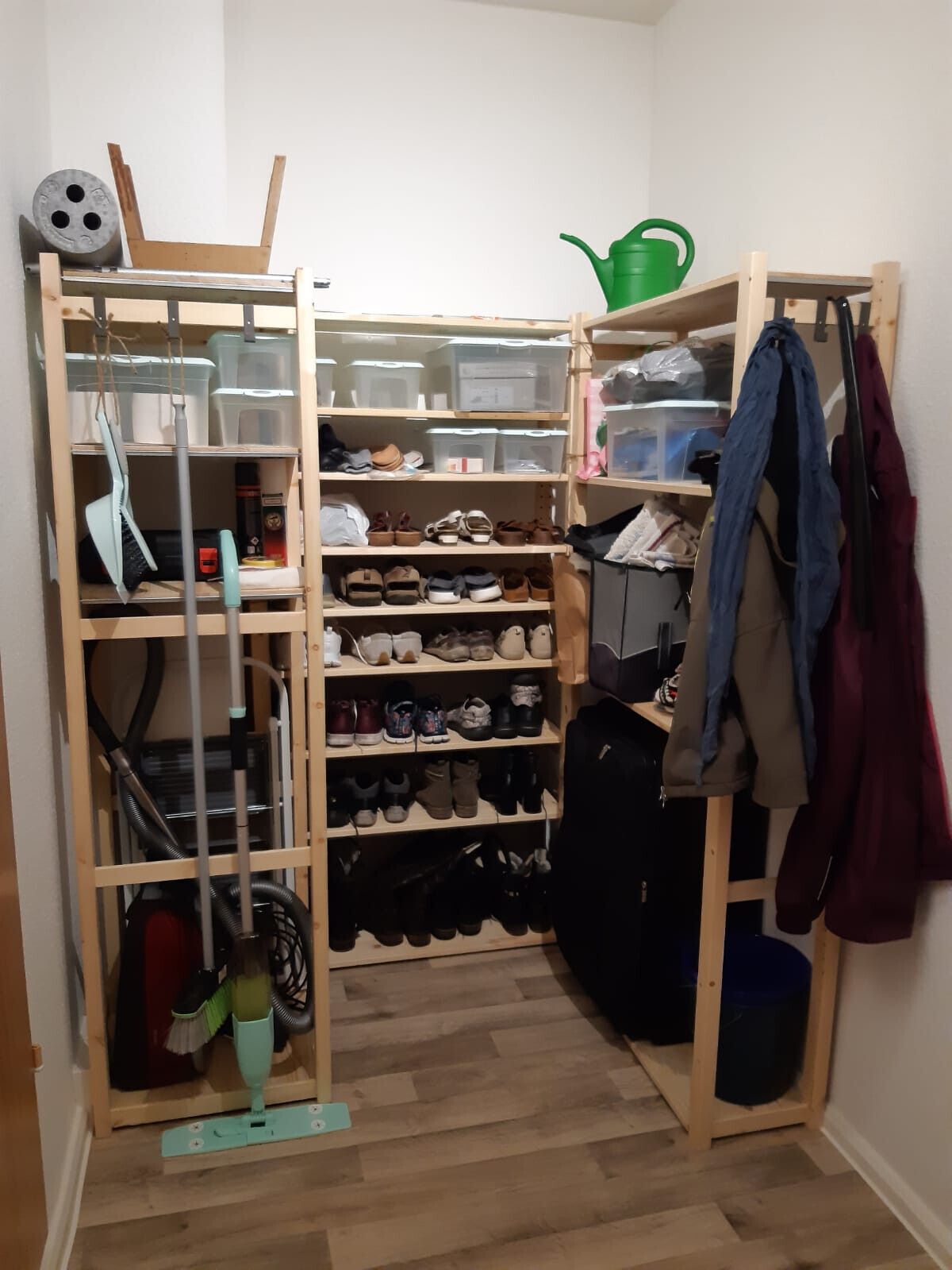 Offener Holzschrank mit Schuhregalen, Aufbewahrungsboxen, Jacken und Reinigungswerkzeug.