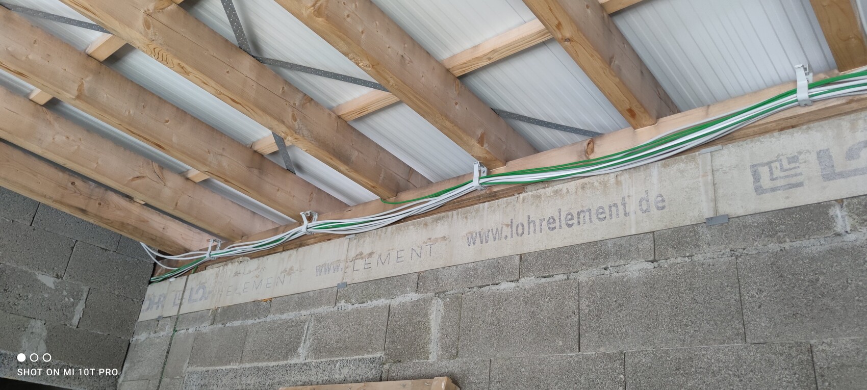 Baustellen-Decke mit Holzbalken, Metallverbindung und grün-weißen Kabelbündeln an einer Betonwand.