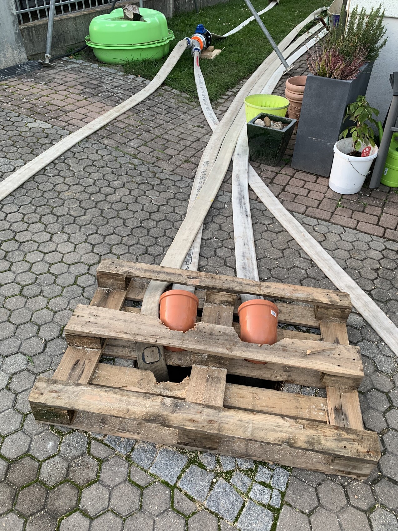 Garten- und Bauutensilien auf gepflastertem Hof: Paletten, Holzlatten, Schläuche, orange Töpfe.