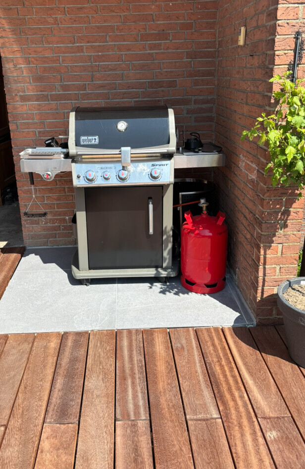 Weber Spirit Gasgrill auf Terrasse neben roter Propanflasche vor Backsteinwand.