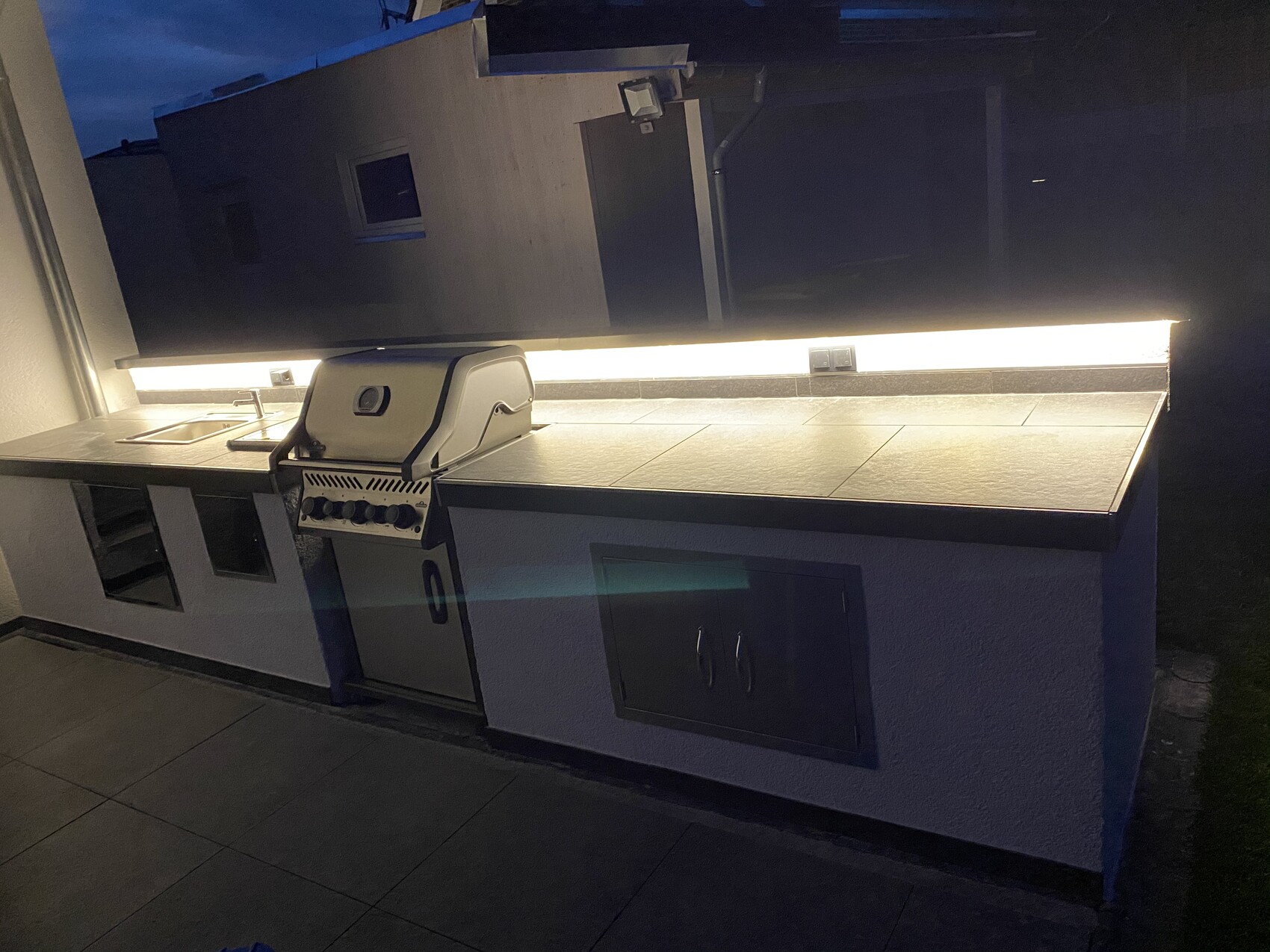 Außenküche mit eingebautem Grill, Spüle und Schränken bei Nacht; LED-Beleuchtung.