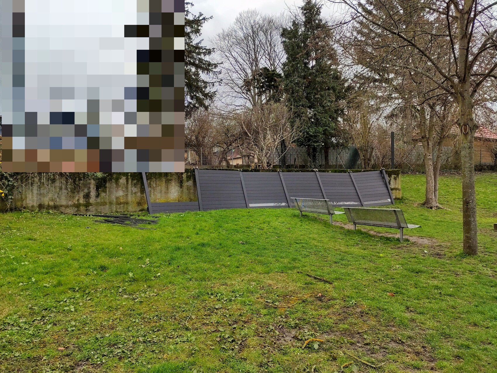 Park mit Gras, zwei Metallbänken, geneigte dunkle Wandpaneele, Bäume im Hintergrund.
