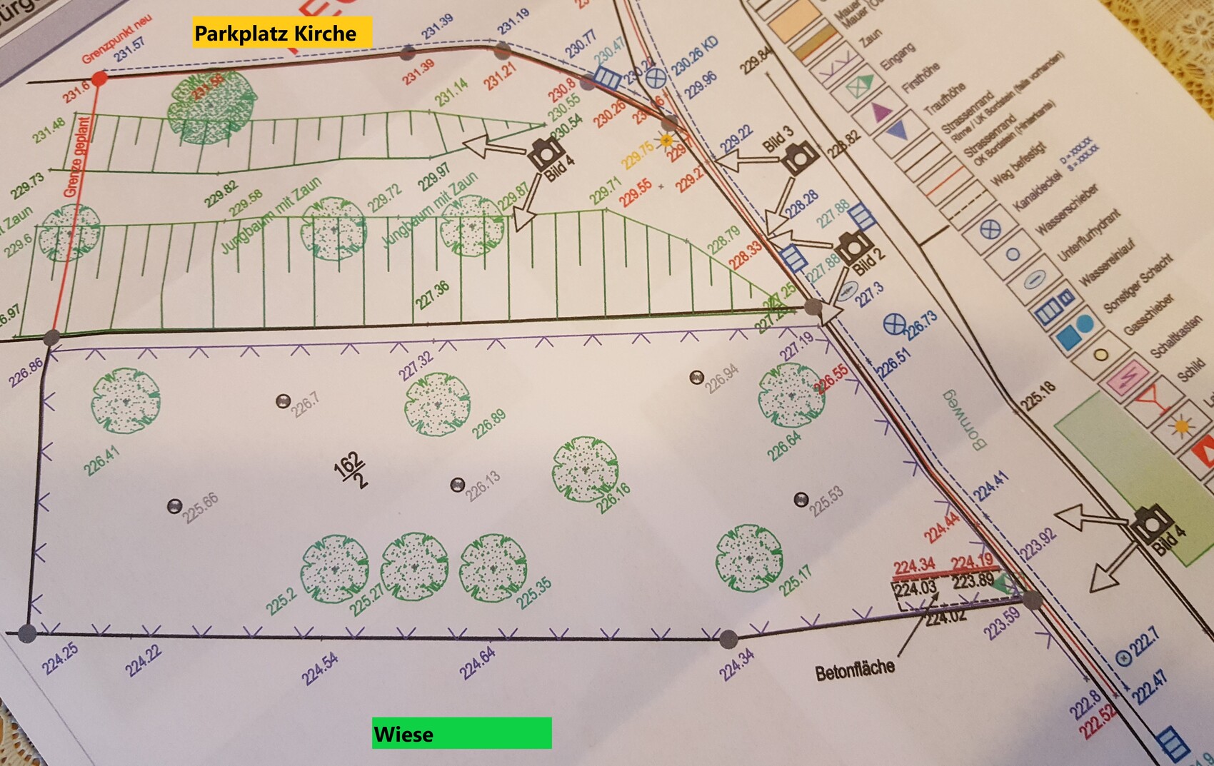 Lageplan: Parkplatz vor der Kirche, grüne Wiese mit Bäumen, Grenzlinien und Straßen.