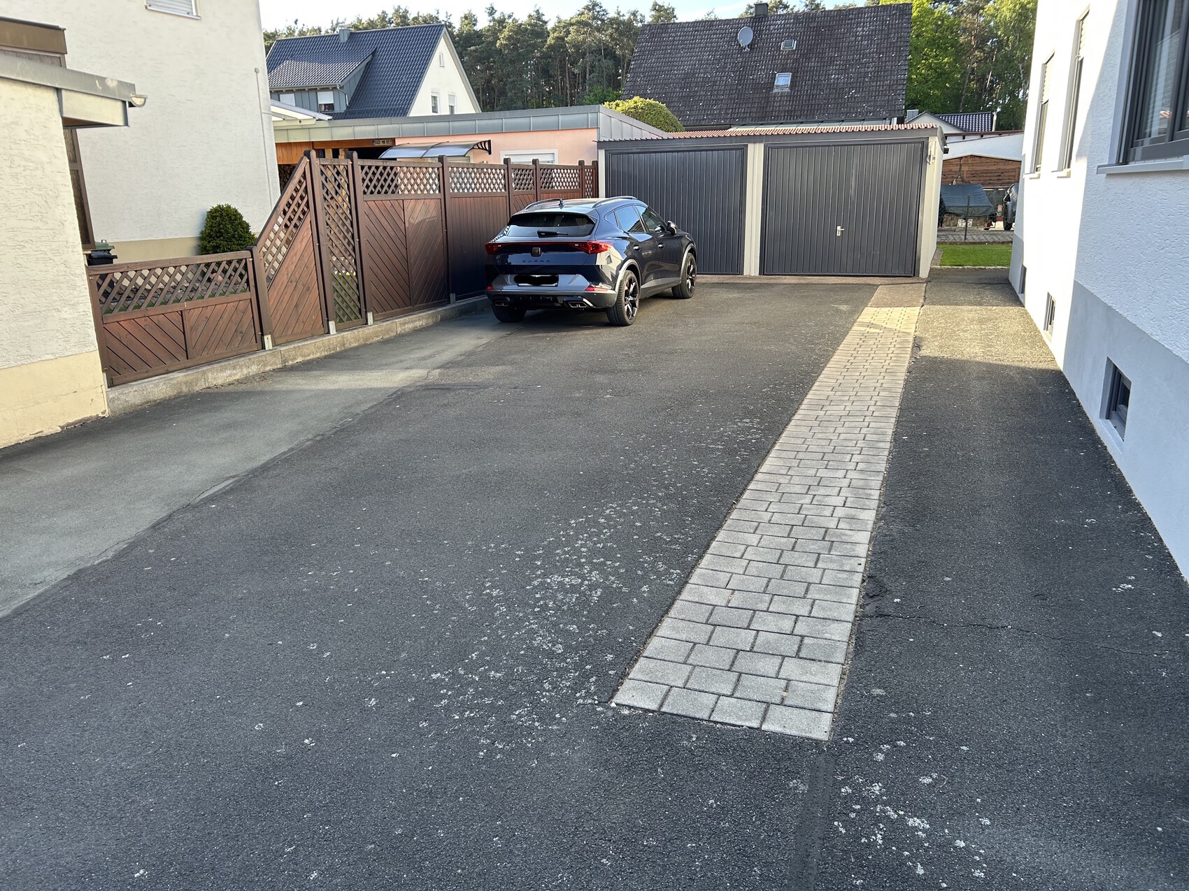 Grauer SUV parkt in der Einfahrt vor zwei Garagentoren; zentrale Pflasterstrecke, Holzzäune links.