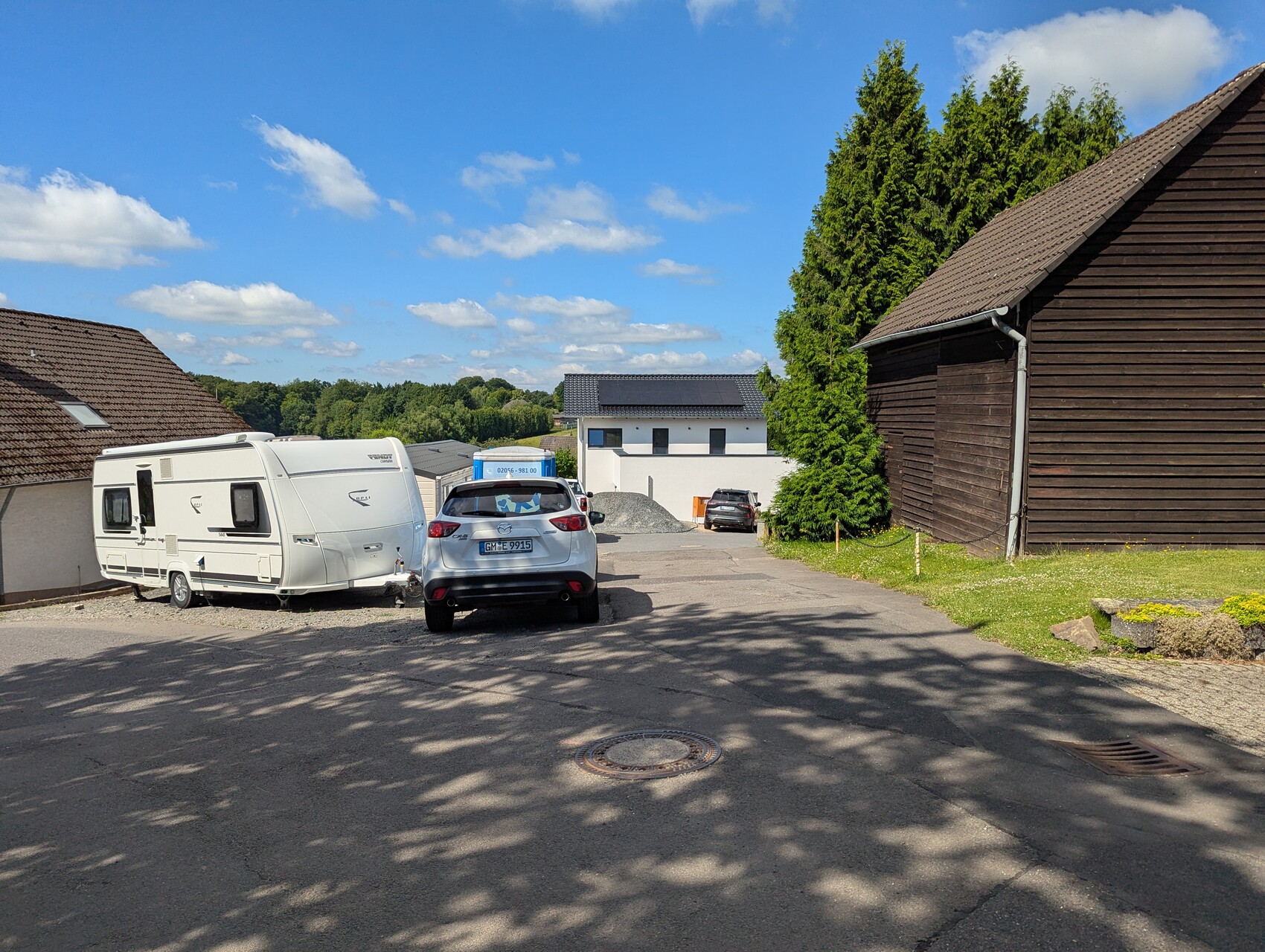 Wohnwagen links, silberner PKW davor, Holzhaus rechts, Bäume und blauer Himmel