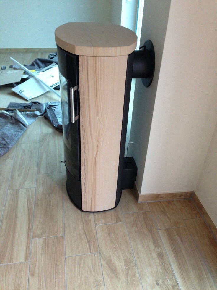 Runder Kaminofen mit Holzverkleidung und Glasfront steht im Zimmer neben der Wand.