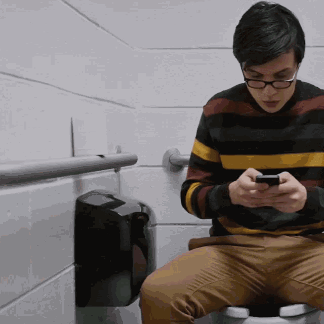Junger Mann mit Brille sitzt auf der Toilette im Badezimmer und tippt auf seinem Smartphone.
