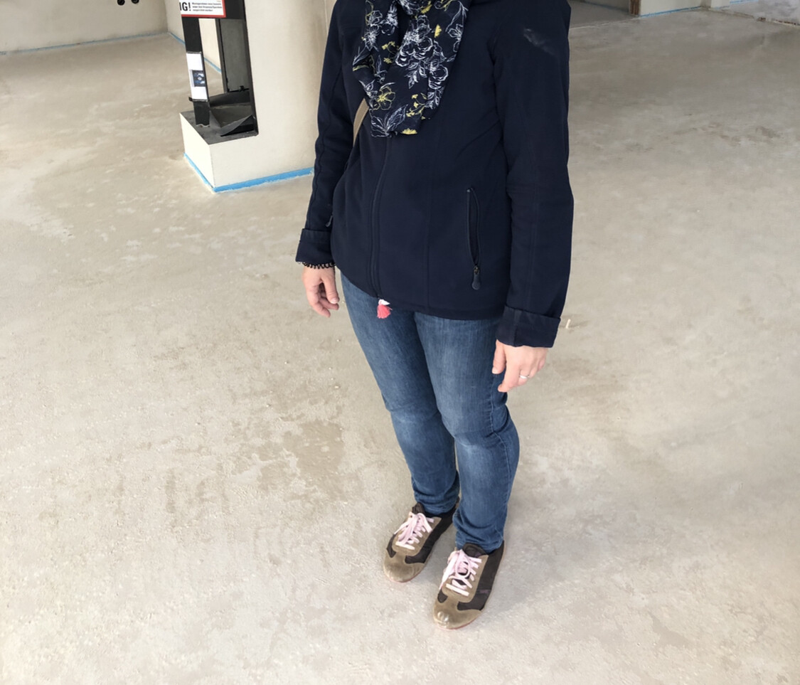 Person steht im Innenraum, dunkelblaue Jacke, gemusterter Schal, Jeans, rosa Sneaker.