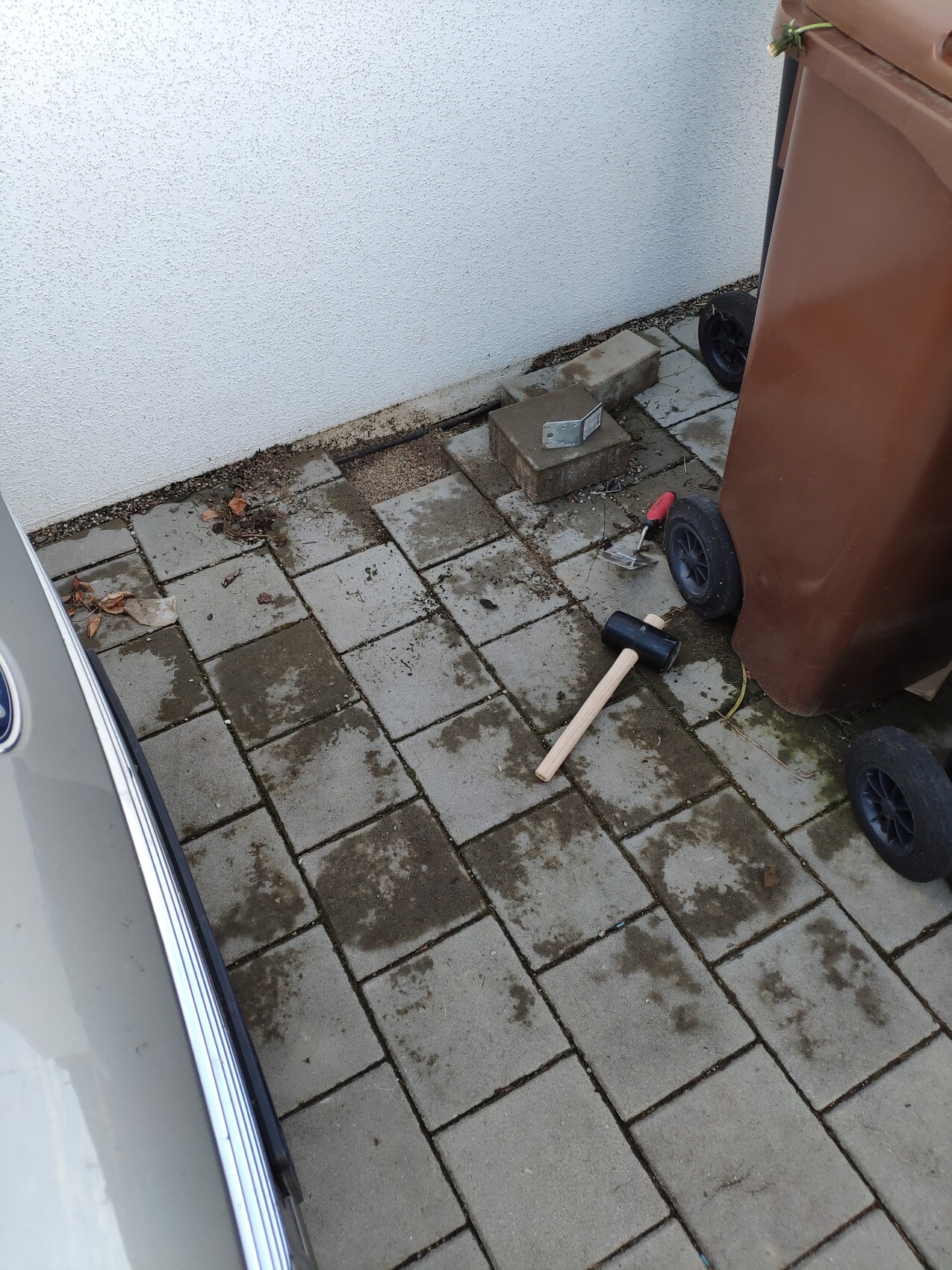 Pflasterpatio neben weißer Wand; brauner Müllcontainer, Steine und Werkzeuge liegen verstreut.