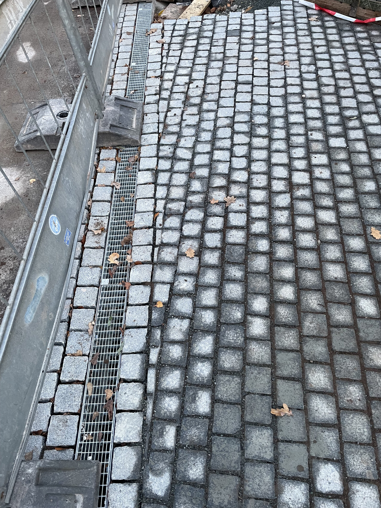 Gepflasterter Vorplatz mit Drainagekanal und Bauzäunen am Rand, Herbstlaub auf Steinen