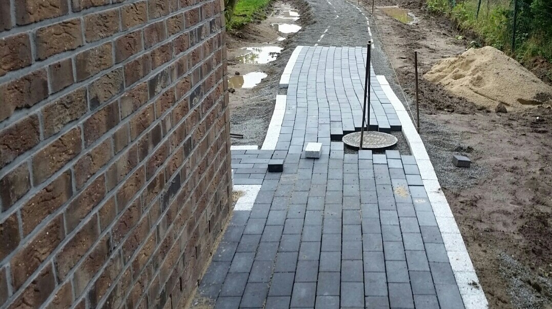 Gehweg aus grauen Pflastersteinen neben Backsteinwand, Runddeckung sichtbar, Sandhaufen; Baustelle