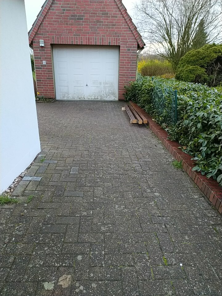 Pflasterweg führt zur Garage mit weißem Tor; Bretter liegen darauf, rechts eine Hecke.