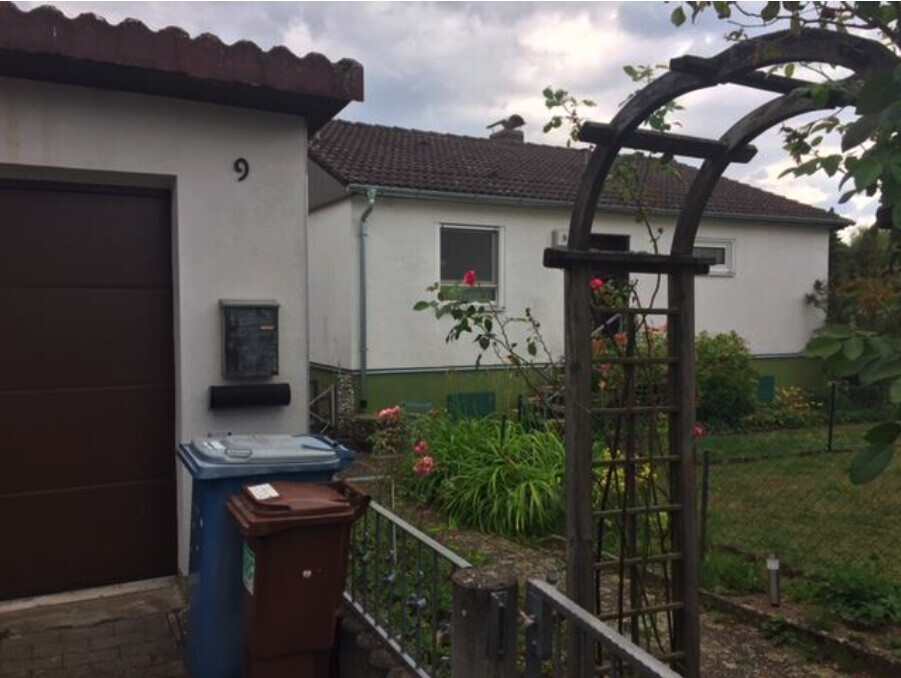 Garten mit Holzbogen-Treillis, Rosen, Mülltonnen neben Garage; weißes Haus dahinter.