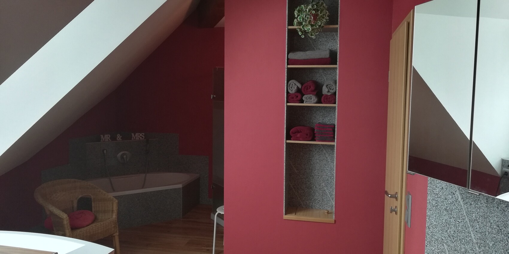 Wellnessbad im Dachgeschoss mit roter Wand, Eckbadewanne, Regal mit Handtüchern und Korbstuhl.