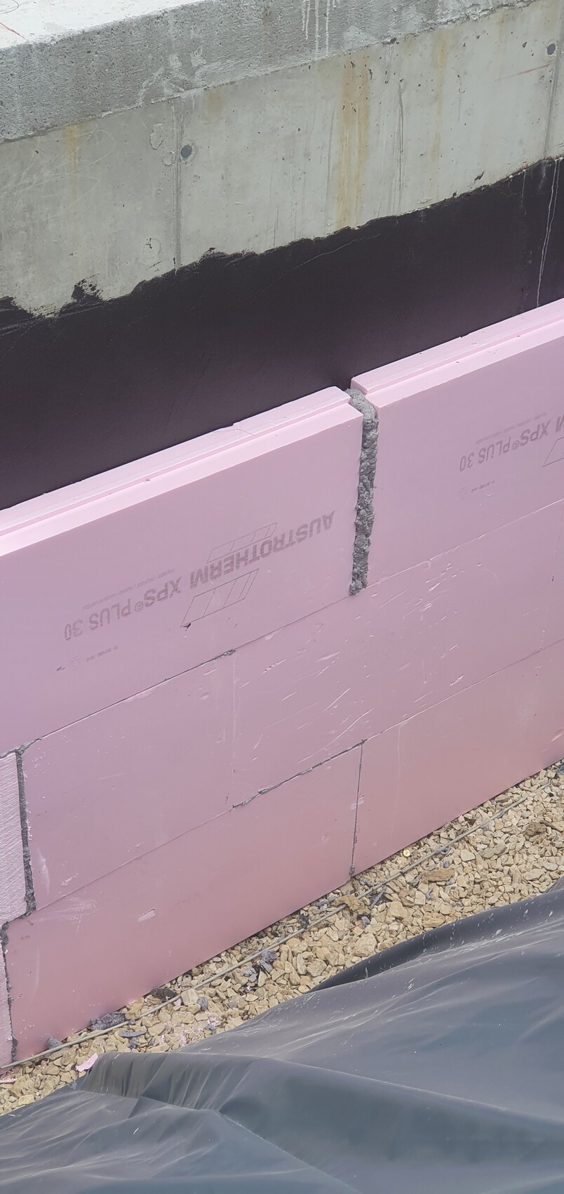 Bauwand: rosa Dämmplatten (Polystyrol) gegen Betonwand; grauer Fugenmörtel; Kiesboden und Baufolie.