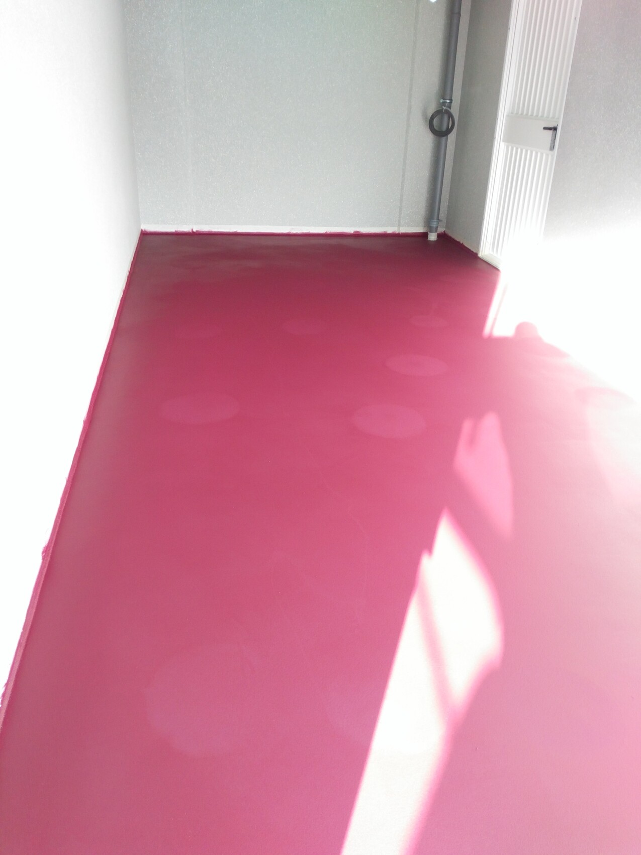 Raum mit pinkem Boden, grauen Wänden, Rohrleitung an der Wand und weißem Heizkörper rechts.