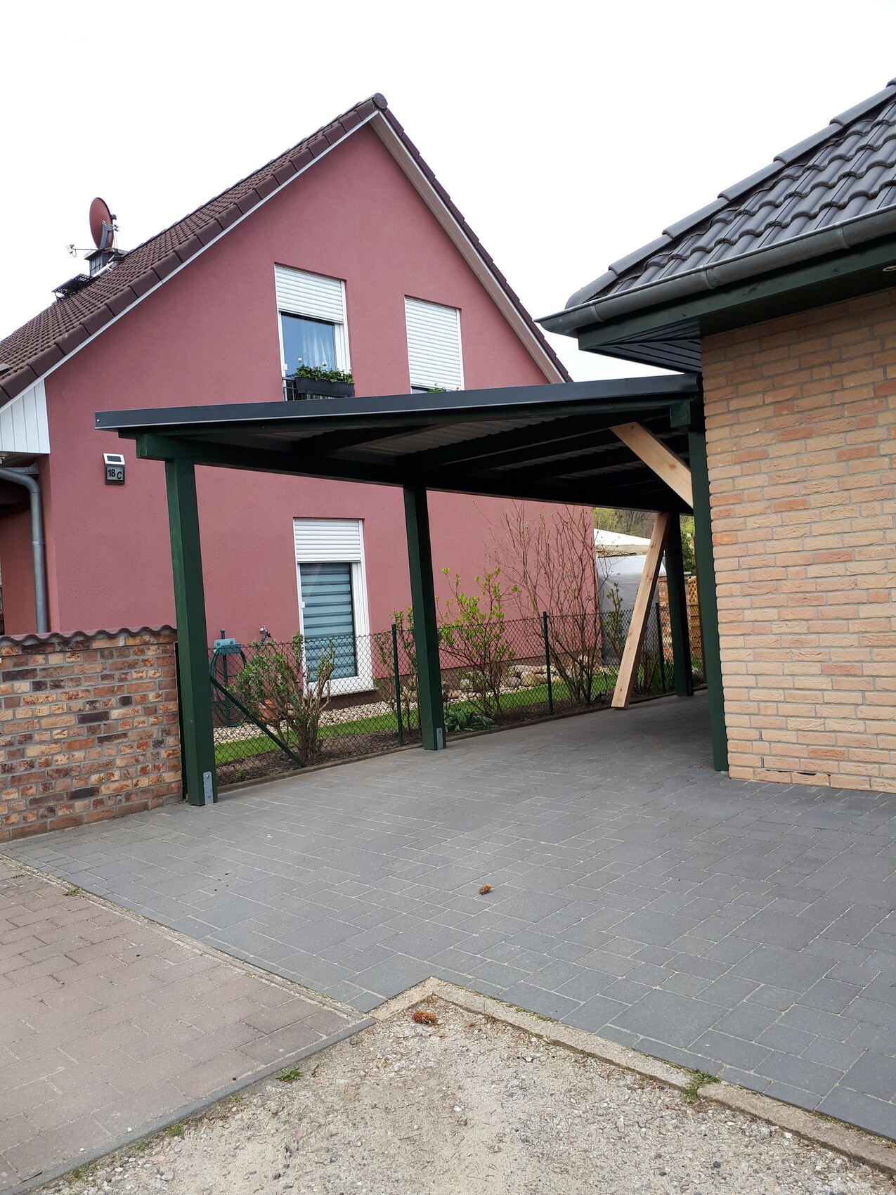 Carport mit grünen Stützsäulen vor pinkem Haus und gepflastertem Hof