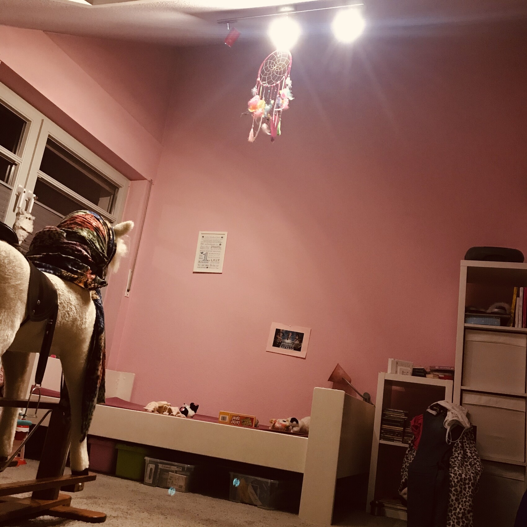Pinkes Kinderzimmer mit Traumfänger unter Deckenleuchte, links Schaukelpferd, rechts Schränke.