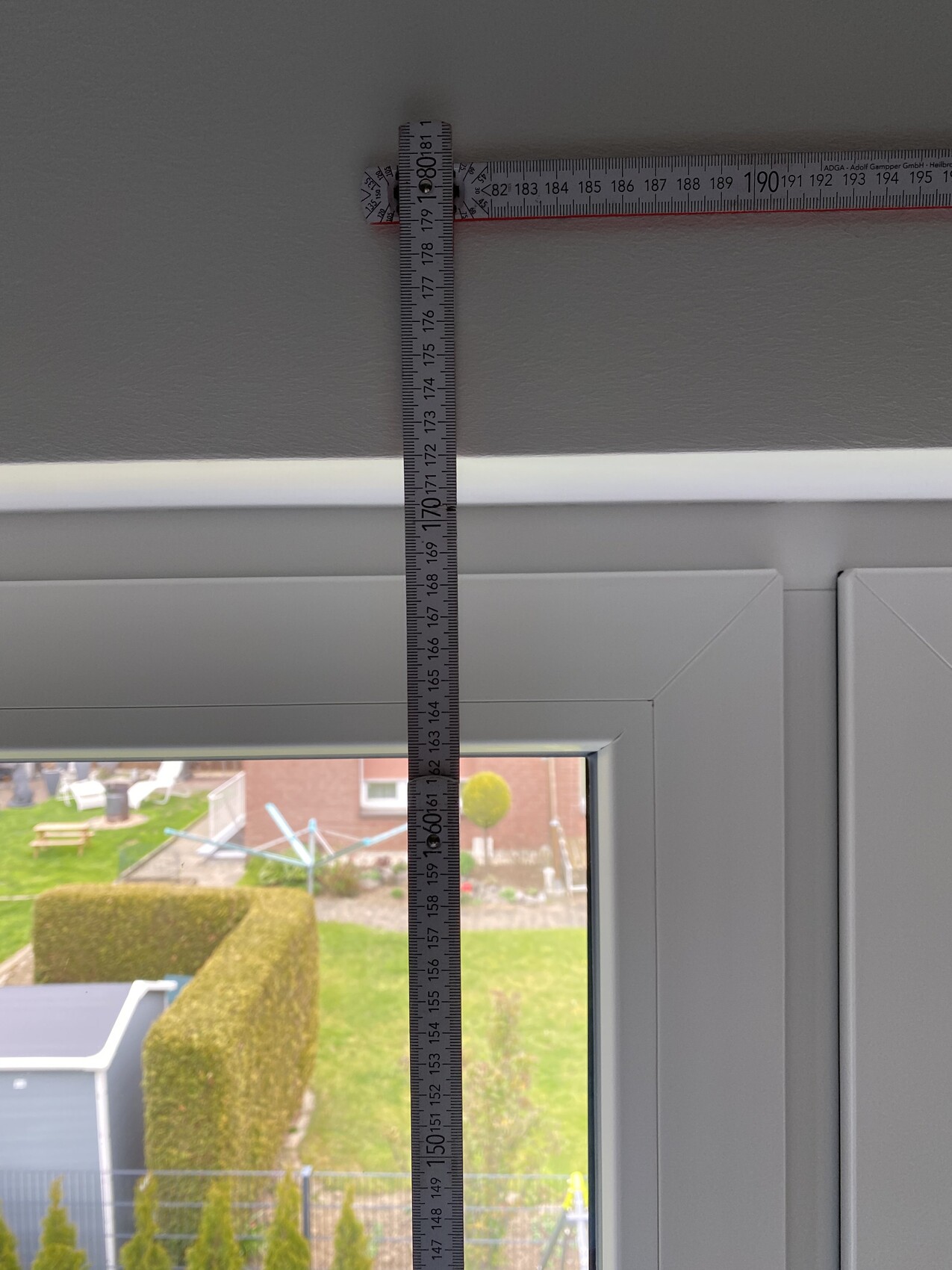 Vertikales Maßband an der Innenwand über einem Fenster, Blick auf den Garten außerhalb.