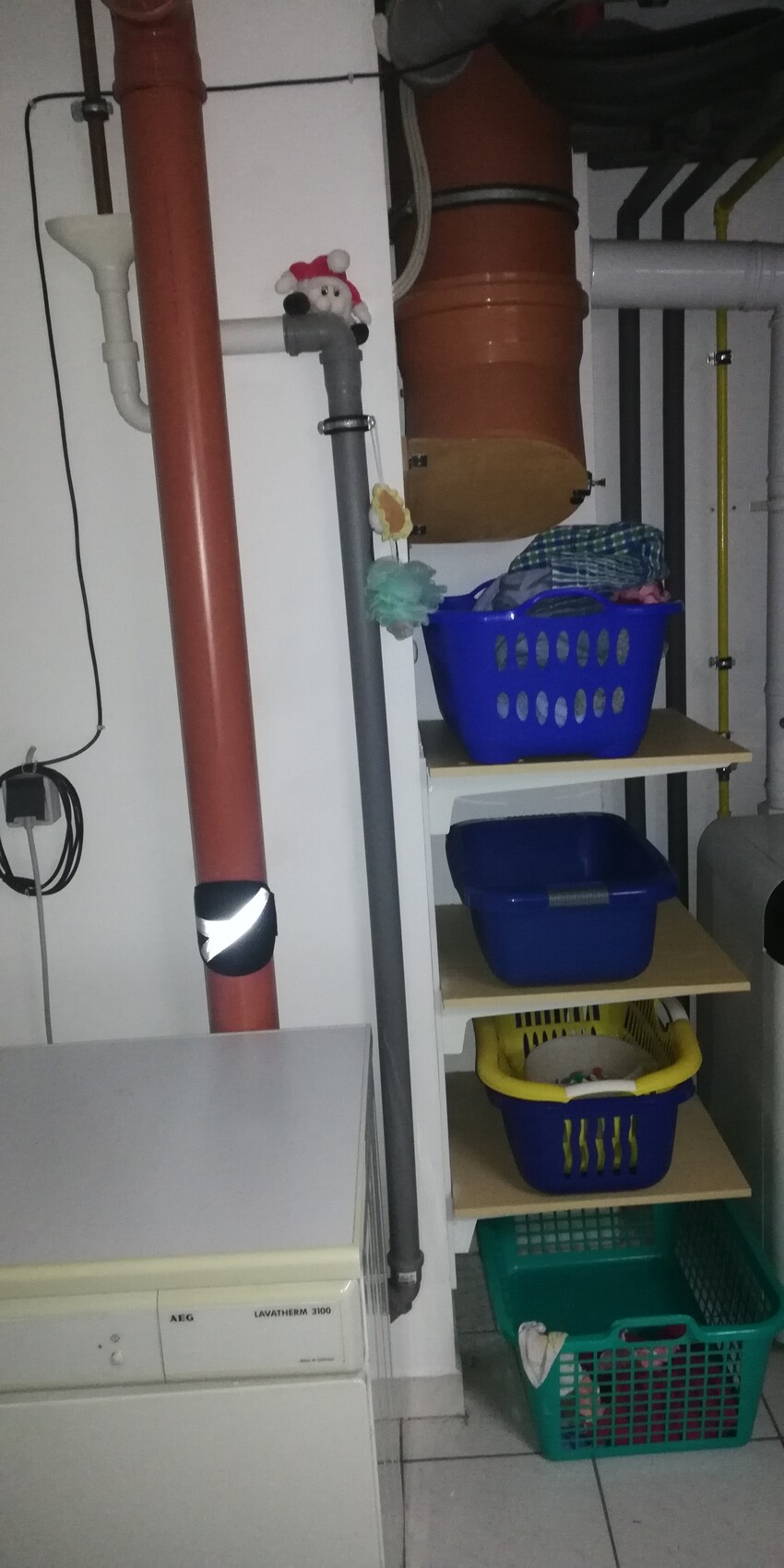 Waschküche/Keller: rote Abflussrohre, Waschmaschine links, Regale mit bunten Wäschekörben, Santa.