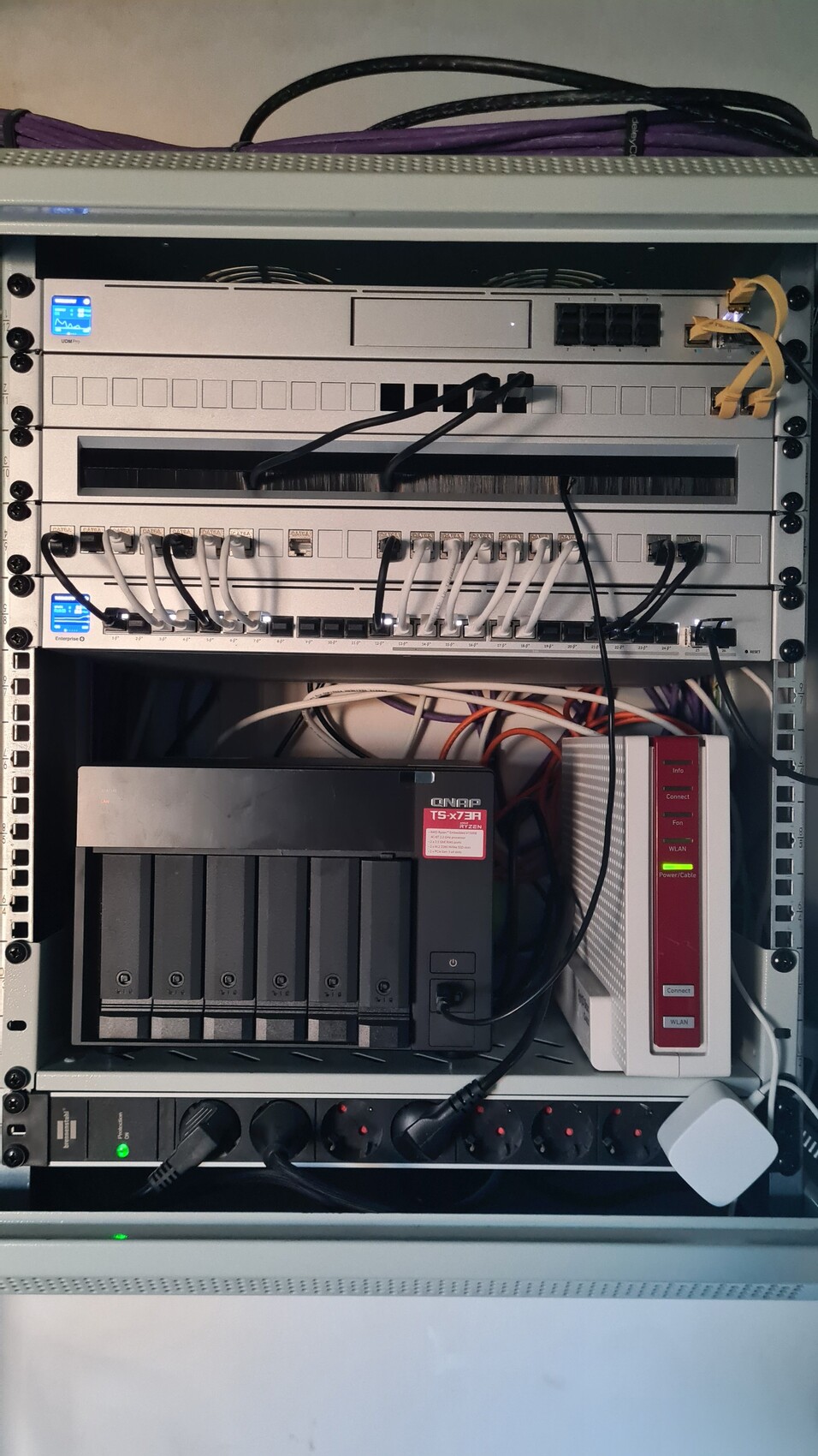 Netzwerkschrank mit QNAP NAS (mehrere Festplatten), Router und vielen Kabeln.