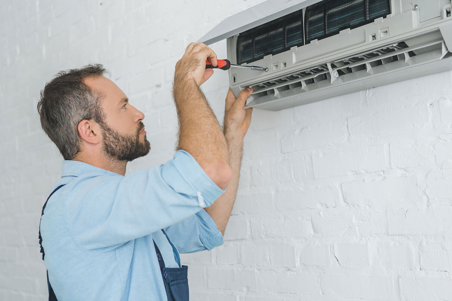 Mann repariert eine Wand-Klimaanlage mit Schraubendreher an einer weißen Ziegelwand.